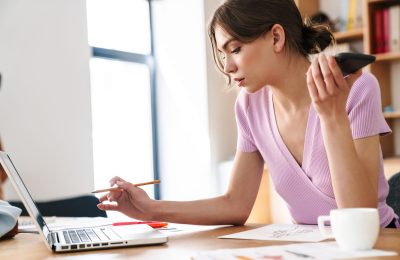 Photo of amazing concentrated woman designer indoors in office using laptop computer while holding mobile phone